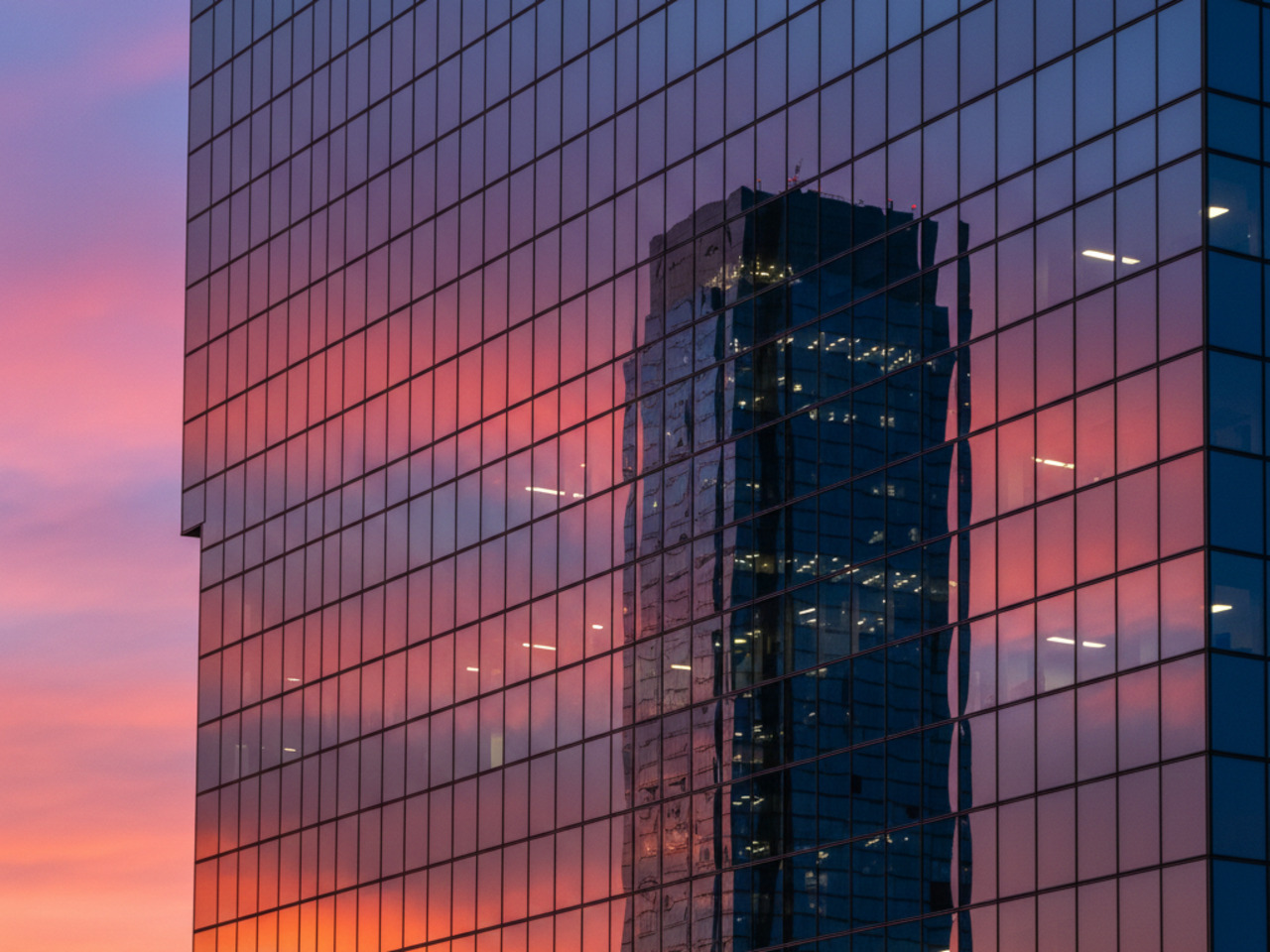 A glass-walled office building reflects a vibrant pink and orange sunset and the dark silhouette of another skyscraper against a twilight sky.