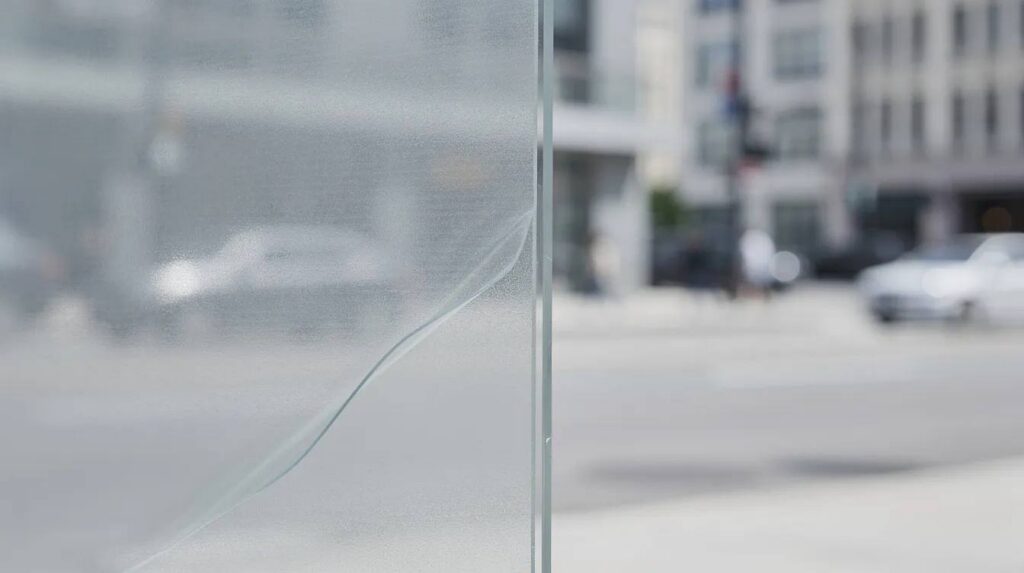 A close-up view of a large commercial office window showcases a clear security window film, designed to hold shattered glass and protect against flying glass shards in the event of explosions. The city street is softly blurred in the background, emphasizing the film's role as a protective barrier for occupants and property.